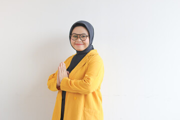 beautiful young asian woman in glasses, hijab and wearing yellow blazer posing namaste greeting hands with smiling face