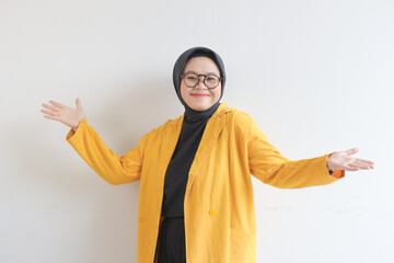 Beautiful young Asian Muslim woman in glasses, hijab and wearing yellow blazer smiling confident and happy with both hands presenting empty space