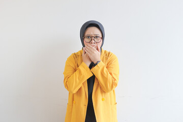 beautiful young Asian Muslim woman, wearing glasses and yellow blazer with shocked expression while covering mouth