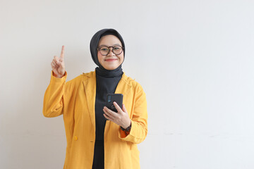 Beautiful young Asian woman in glasses, hijab and wearing a yellow blazer is holding a mobile phone with a smiling face