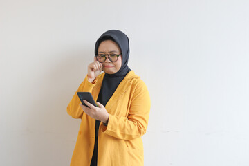 beautiful young asian woman in glasses, hijab and wearing yellow blazer looking at her mobile phone with sullen face