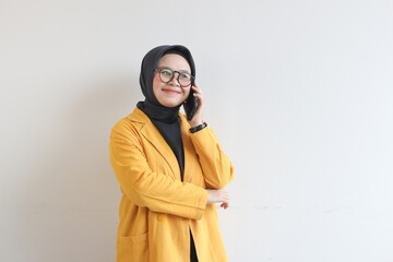 portrait of beautiful asian woman in hijab, glasses and wearing yellow blazer making phone call and smiling