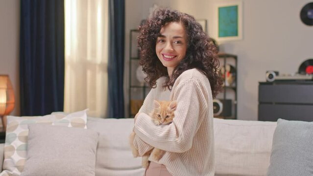 Zooming in on Caucasian woman holding kitten in her hands and petting it with love. Hugging red cat while looking at camera. Enjoying spending time with her domestic pet. Cheerfully smiling on camera.