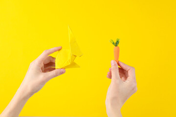 Hand holding a plastic miniature carrot and origami rabbit on a yellow background. Easter concept