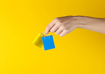 Woman's hand holds yellow and blue miniature shopping bag on yellow background. Sale, shopping...