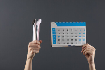 Hands hold toothbrushes and a calendar on a dark gray background. Dental care, visit to the dentist
