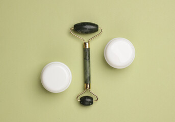 Massage jade roller and cream jars on green background. Beauty concept