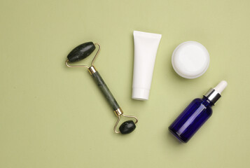 Massage jade roller and serum bottle, cream tube and jar on green background. Beauty concept. Flat lay