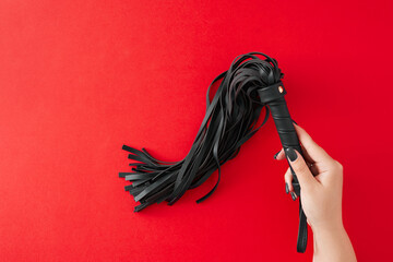 Ignite desires with carefully selected BDSM toys. First person top view photo of female hand holding leather whip on red background with empty space for text or advert - obrazy, fototapety, plakaty