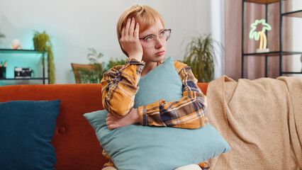 Sad lonely young woman sitting at home room looks pensive thinks over life concerns or unrequited...