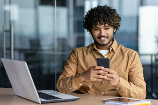 Successful young businessman inside office at workplace, man typing message and browsing social media, hispanic smiling sitting holding phone, browsing online sites.