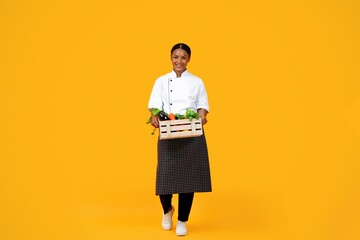Black Chef Woman Carrying Crate With Vegetables While Walking On Yellow Background