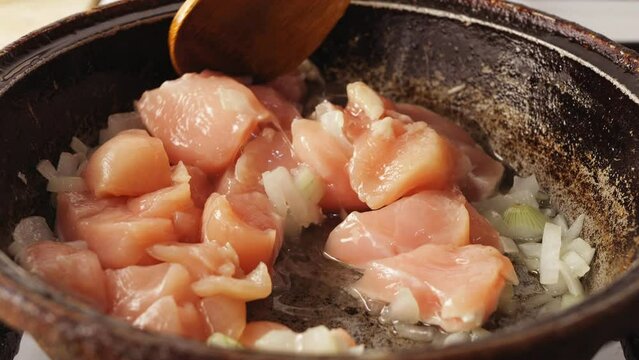 Pieces of chicken fried in a cast-iron skillet in oil are mixed with a wooden spatula. Close-up