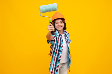 Child builder in hard hat helmet. Teenage girl painter with painting brush tool or paint roller. Teenager worker isolated on yellow background. Kids renovation concept.