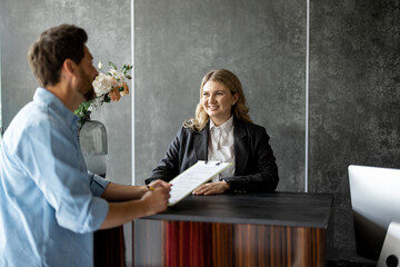 Young medical receptionist talks with a male client and gives some paper to sign at the reception...