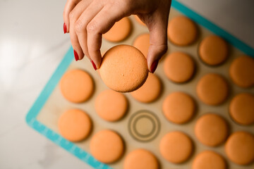 Hand of woman confectioner holds freshly baked half of dessert macaron above silicone baking sheet.