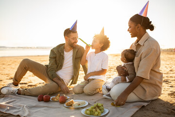Diverse family of four sitting on blanket on the beach at sunset, wearing party hands and blowing whistles - Powered by Adobe