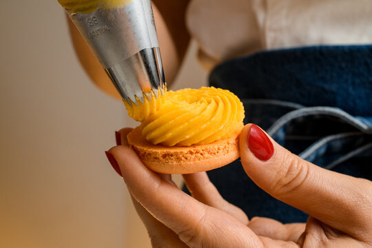 Closeup view on woman chef with confectionery bag in hands squeezing orange cream to fresh macarons halves