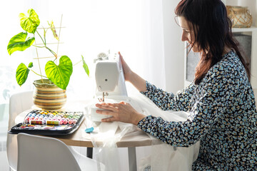 A woman sews tulle on an electric sewing machine in a white modern interior of a house with large...