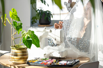 Woman thinking, dreaming and sews tulle on an electric sewing machine in a white modern interior of a house with large windows, house plants. Comfort in house, housewife's hobby