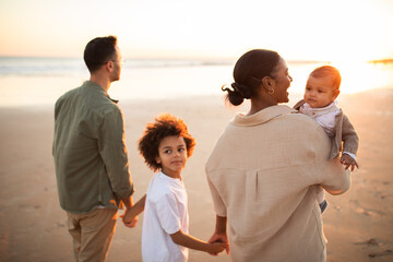 Rear view of black mother and father walking by seaside with two kids, holding hands, enjoying...