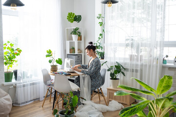 A woman sews tulle on an electric sewing machine in a white modern interior of a house with large windows, house plants. Comfort in the house, a housewife's hobby