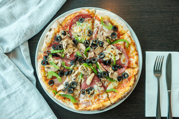 Delicious pizza with mushrooms, capsicum, pepper, black olives, ham, cheese and salami on a wooden table with cutlery and napkin
