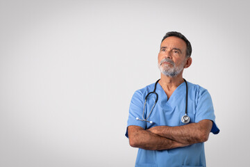 Serious pensive caucasian senior doctor in blue uniform with crossed arms on chest looking at free space