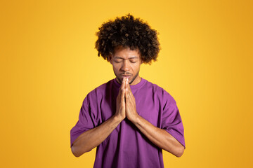 Fototapeta na wymiar Calm serious black adult curly man with closed eyes in purple t-shirt prays with hands
