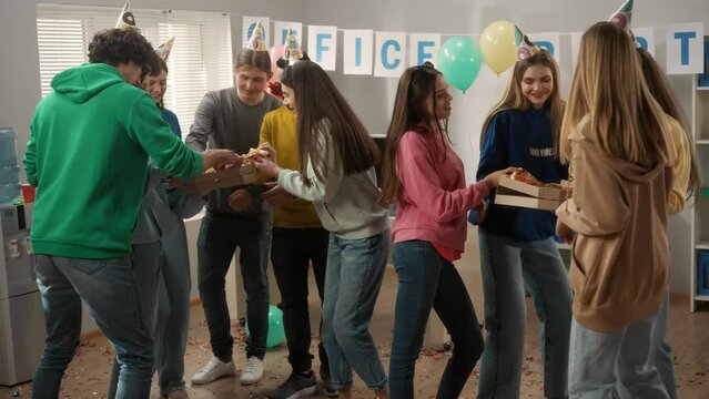 A group of young people in party hats and accessories are dancing in the office at a party. Modern office workers dance and eat pizza from boxes held by their colleagues. Party, birthday, office party