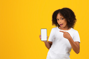 Glad shocked curly teenager black schoolgirl in white t-shirt pointing finger at smartphone with...