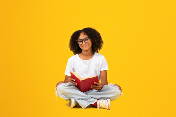 Cheerful smart curly teenager black schoolgirl in white t-shirt and glasses reads book