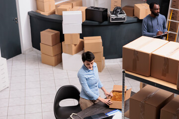 Top view of employee standing at desk in warehouse while preparing client order putting clothes in carton box after checking delivery detalies. Diverse team working in distribution center