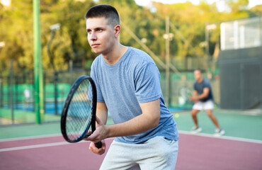 Young man in shorts and t-shirt playing tennis on court. Racket sport training outdoors.