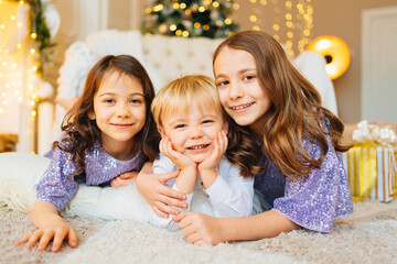 two girls and a little boy lie on the floor in a room with Christmas decor.