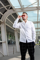a handsome man nicely and stylishly dressed in pink glasses stands at a bus stop