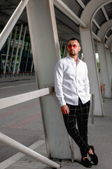 a handsome man nicely and stylishly dressed in pink glasses stands at a bus stop