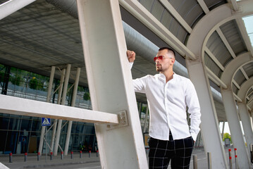 a handsome man nicely and stylishly dressed in pink glasses stands at a bus stop