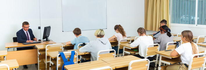 Back view of group of teenage students studying in classroom with male teacher..