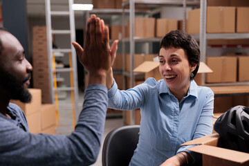 Smiling employees doing high five in storehouse after finishing putting helmet order for client in...