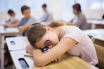 Exhausted teen girl sleeping at desk in classroom during lesson with blurred classmates in background