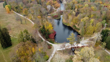 Beautiful scenery view of an autumn park with trees with yellow fallen leaves, lakes,waterfall, architecture, glades and people walking dirt paths on autumn day. Flying over the autumn park. Top view.