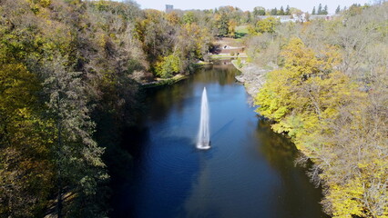 Obraz premium Fountain with rainbow in middle of lake between trees with yellow leaves in park on sunny autumn day. Decorative fountain in center of lake. Natural park arboretum in autumn. Natural landscape. Aerial