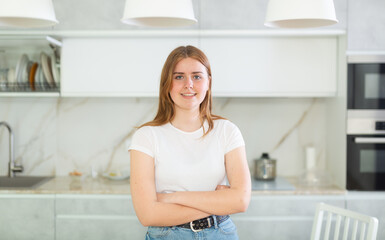 Portrait of positive girl teenager in casual wear smiling at camera while standing with crossed hands in cozy kitchen