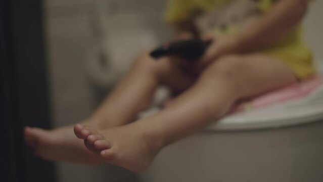 little baby child sit on toilet on pink child seat holding black TV remote hands. caucasian toddler boy yellow t-shirt learning use toilet. hygiene education preschooler health care concept. WC kid 