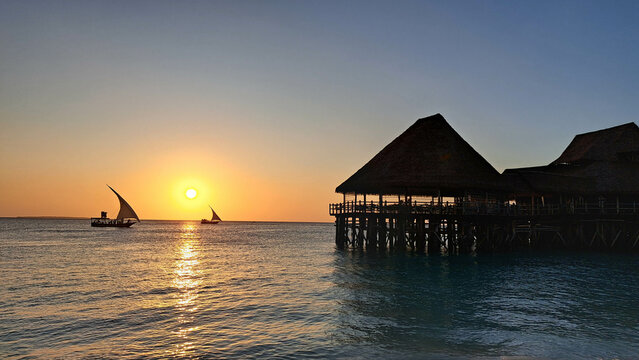 Kendwa, Zanzibar Island, Tanzania dhow boats sailing and a wooden thatched bungalow pier against the setting sun and cloudy sky.