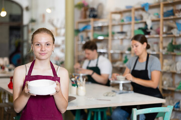 Girl teenager student in apron posing with ceramic bowl in her hands in ceramic workshop