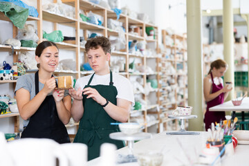 Young woman teacher in apron examines ceramic cup made by teenage boy student