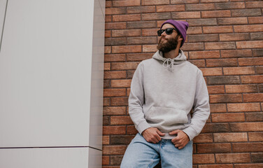 City portrait of handsome hipster guy with beard wearing gray blank hoodie or sweatshirt and hat...