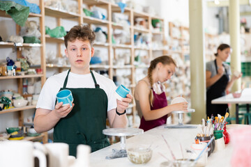 Guy teenager student in apron posing with ceramic cups in his hands in ceramic workshop
